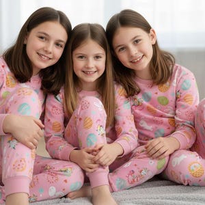 May include: Three people wearing matching pink pyjamas with an Easter-themed pattern. The pyjamas feature illustrations of Easter eggs, bunnies, and baskets. They are sitting on a grey blanket.