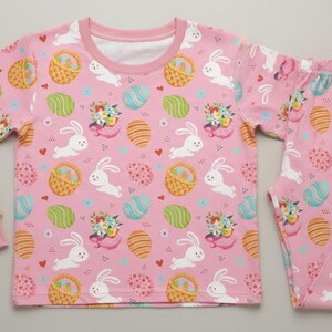 May include: A pink children's pyjama set featuring a pattern of white rabbits, Easter eggs, and baskets. The long-sleeved top and trousers are made of a soft material. The set is designed for children.