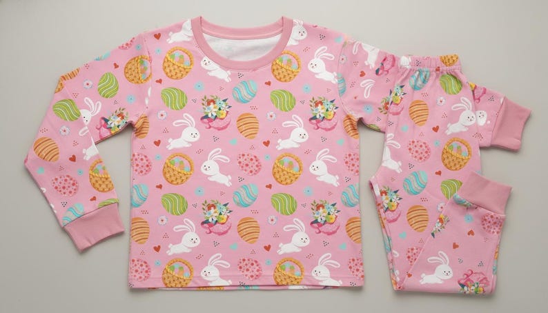 May include: A pink children's pyjama set featuring a pattern of white rabbits, Easter eggs, and baskets. The long-sleeved top and trousers are made of a soft material. The set is designed for children.