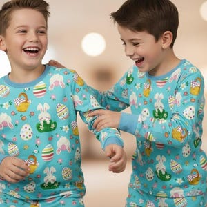 May include: Two children wearing matching light blue Easter-themed pajamas. The pajamas feature a pattern of Easter eggs, bunnies, and baskets. The long-sleeved tops have blue cuffs and necklines. The children are smiling and laughing.