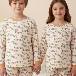 K&ouml;nnte beinhalten: Passende cremefarbene Oster-Schlafanz&uuml;ge mit langen &Auml;rmeln und Hosen. Die Schlafanz&uuml;ge sind mit dem Wort "EASTER" in verschiedenen Farben gemustert. Die Kinder halten sich an den H&auml;nden.