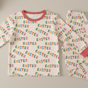 K&ouml;nnte beinhalten: Ein cremefarbenes Pyjama-Set mit dem Wort "EASTER" in verschiedenen Farben bedruckt. Das lang&auml;rmlige Oberteil und die Hose haben korallenfarbene B&uuml;ndchen an den Manschetten und am Halsausschnitt. Ein festliches und bequemes Schlafanzug-Set.