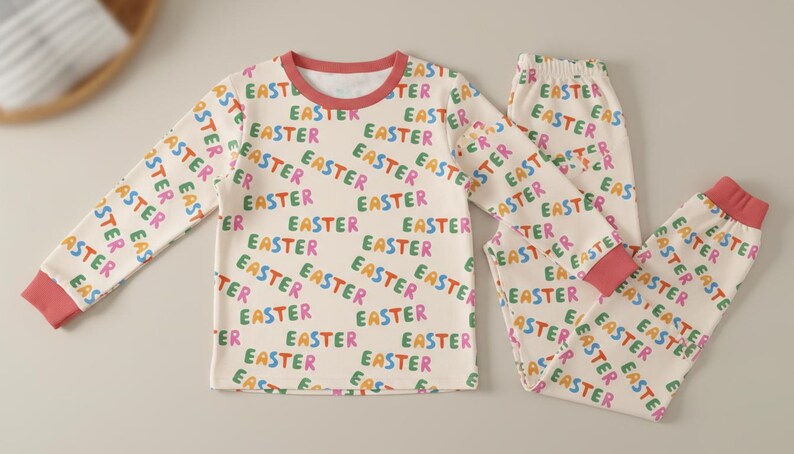 K&ouml;nnte beinhalten: Ein cremefarbenes Pyjama-Set mit dem Wort "EASTER" in verschiedenen Farben bedruckt. Das lang&auml;rmlige Oberteil und die Hose haben korallenfarbene B&uuml;ndchen an den Manschetten und am Halsausschnitt. Ein festliches und bequemes Schlafanzug-Set.