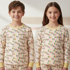 K&ouml;nnte beinhalten: Zwei Kinder in passenden cremefarbenen Pyjamas mit dem Wort "EASTER" in verschiedenen Farben bedruckt. Die Pyjamas haben lange &Auml;rmel und Hosen. Die Kinder l&auml;cheln und stehen vor einem Bett.