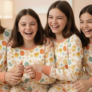 May include: Four people wearing matching Easter-themed pyjamas. The cream-coloured pyjamas have orange trim and a pattern of Easter eggs, chicks, and bunnies. The people are smiling and laughing.