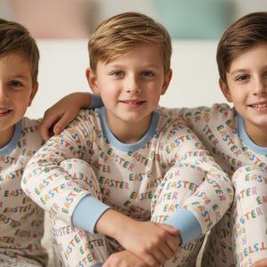 K&ouml;nnte beinhalten: Drei junge Menschen in passenden Pyjamas mit dem Wort "EASTER" in verschiedenen Farben. Die Pyjamas haben hellblaue B&uuml;ndchen und Ausschnitte. Die Personen l&auml;cheln und sitzen zusammen.