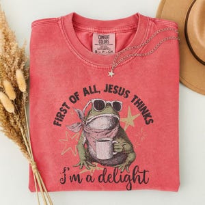 May include: A coral-colored crewneck t-shirt featuring a graphic of a green frog wearing sunglasses and a bandana while holding a coffee mug. The text reads, First of all, Jesus thinks I'm a delight. The shirt has a Comfort Colors brand label.
