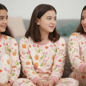 Puede incluir: Tres j&oacute;venes con pijamas a juego. Los pijamas son de color rosa claro con un estampado de flores, corazones y otras formas. La parte superior de manga larga y los pantalones tienen ribetes rosas.