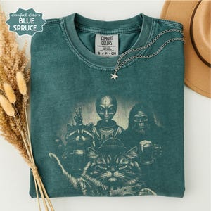 May include: A blue spruce coloured t-shirt featuring a graphic print of an alien, a raccoon, a sasquatch, and a cat, all holding mugs. The shirt has a crew neckline and a Comfort Colors brand label on the inside collar. A silver star necklace is placed on top.