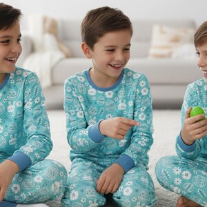May include: Three children wearing matching teal pajamas with an Easter-themed print of eggs, bunnies, and flowers. The pajamas have blue trim and long sleeves. The children are smiling and holding Easter eggs.
