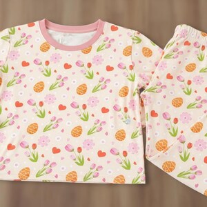 Puede incluir: Un conjunto de pijama de dos piezas con fondo crema y un estampado repetido de huevos de Pascua naranjas, flores rosas y corazones rojos. La parte superior de manga larga y los pantalones tienen ribetes rosas.