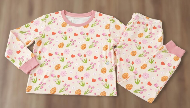 Puede incluir: Un conjunto de pijama de dos piezas con fondo crema y un estampado repetido de huevos de Pascua naranjas, flores rosas y corazones rojos. La parte superior de manga larga y los pantalones tienen ribetes rosas.