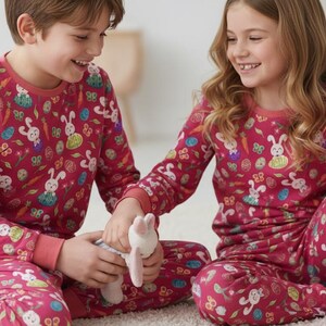 May include: Two children wearing matching red pyjamas with a bunny and Easter egg pattern. The pyjamas have long sleeves and trousers. They are sitting on a white rug and holding a stuffed bunny.