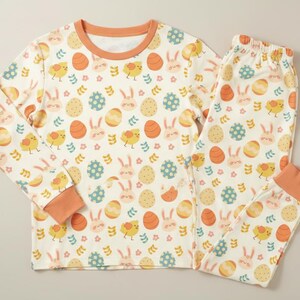 May include: A cream-coloured two-piece pyjama set featuring an Easter-themed pattern. The pattern includes orange eggs, yellow chicks, and light blue and pink bunnies. The long-sleeved top and trousers have orange trim.
