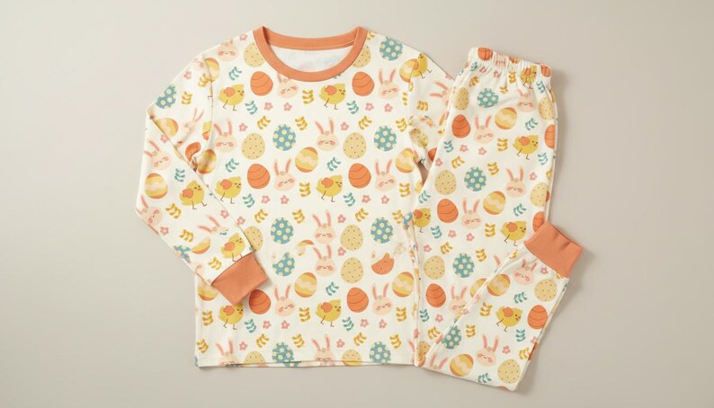 May include: A cream-coloured two-piece pyjama set featuring an Easter-themed pattern. The pattern includes orange eggs, yellow chicks, and light blue and pink bunnies. The long-sleeved top and trousers have orange trim.
