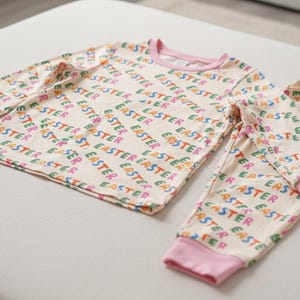 K&ouml;nnte beinhalten: Ein zweiteiliger Schlafanzug mit cremefarbenem Oberteil und Hose. Das Set ist mit dem Wort "EASTER" in verschiedenen Farben bedruckt. Die B&uuml;ndchen und der Halsausschnitt sind rosa.