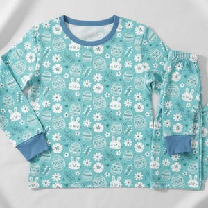 May include: A two-piece pajama set in a light teal color, featuring a repeating pattern of Easter eggs, bunnies, and flowers. The long-sleeved top and pants have blue cuffs and trim. The number "2" is in the top left corner.
