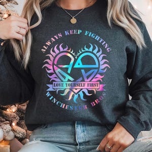 Może przedstawiać: Szara bluza z holograficznym wzorem serca. Serce otoczone jest promieniami słońca, a na niej widnieje napis "Always Keep Fighting Love Yourself First Winchester Bros".