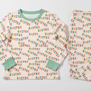 K&ouml;nnte beinhalten: Cremefarbenes Oster-Pyjama-Set mit einem lang&auml;rmligen Oberteil und einer Hose. Der Pyjama ist mit dem Wort "EASTER" in verschiedenen Farben bedeckt. Die B&uuml;ndchen und der Halsausschnitt sind mintgr&uuml;n.