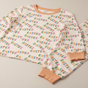 K&ouml;nnte beinhalten: Cremefarbenes Pyjama-Set mit dem Wort "EASTER" in verschiedenen Farben wiederholt gedruckt. Das lang&auml;rmlige Oberteil und die Hose haben pfirsichfarbene B&uuml;ndchen und Bes&auml;tze. Ein festliches Outfit f&uuml;r Ostern.