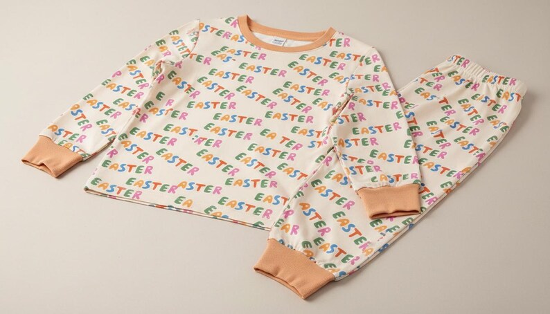 K&ouml;nnte beinhalten: Cremefarbenes Pyjama-Set mit dem Wort "EASTER" in verschiedenen Farben wiederholt gedruckt. Das lang&auml;rmlige Oberteil und die Hose haben pfirsichfarbene B&uuml;ndchen und Bes&auml;tze. Ein festliches Outfit f&uuml;r Ostern.