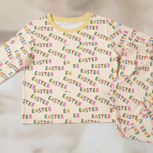 K&ouml;nnte beinhalten: Cremefarbenes Ostern-Pyjama-Set mit einem lang&auml;rmeligen Oberteil und einer Hose. Das Pyjama ist mit dem Wort "EASTER" in verschiedenen Farben bedruckt. Die B&uuml;ndchen und der Halsausschnitt sind gelb.