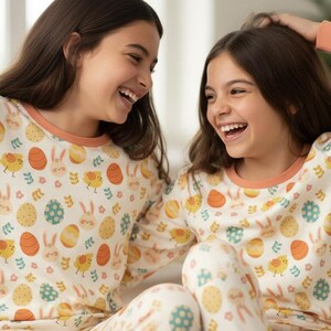 May include: Two young people wearing matching Easter-themed pyjamas, smiling and laughing. The pyjamas are cream-coloured with a pattern of Easter eggs, chicks, and bunnies. The pyjamas have orange trim on the neckline and cuffs.