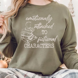 May include: Olive green sweatshirt with the text "emotionally attached to fictional characters" in white script. The graphic includes a stack of books and a coffee cup. The sweatshirt is a comfortable, casual style.