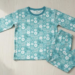 May include: Teal-colored pajama set with a repeating pattern of white Easter eggs, bunnies, and flowers. The long-sleeved top and pants have teal cuffs and a white neckline. The set is made of a soft, comfortable material.