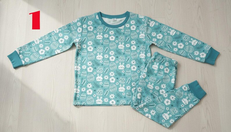 May include: Teal-colored pajama set with a repeating pattern of white Easter eggs, bunnies, and flowers. The long-sleeved top and pants have teal cuffs and a white neckline. The set is made of a soft, comfortable material.
