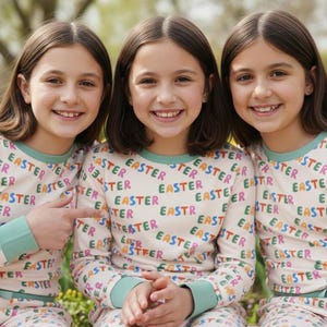 K&ouml;nnte beinhalten: Drei junge Menschen tragen passende Pyjamas mit dem Wort "EASTER" in bunten Buchstaben. Sie l&auml;cheln und posieren im Freien, mit einem Hintergrund aus Blumen und Gr&uuml;n.