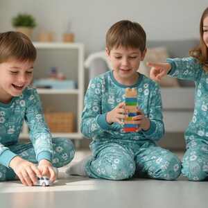 May include: Three children wearing matching teal pajamas with a floral and bunny print are playing on the floor. One child holds a toy car, another a wooden block tower, and the third points and laughs.