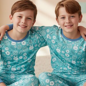 May include: Two children wearing matching teal pajamas with an Easter-themed pattern of eggs, bunnies, and flowers. The pajamas have blue trim and long sleeves. The children are smiling and sitting on a light-colored rug.