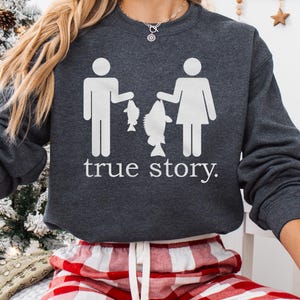 Puede incluir: Una sudadera gris oscuro con un gráfico blanco de dos figuras de palo sosteniendo peces. El texto "true story." está impreso debajo de las figuras.