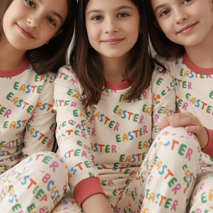 K&ouml;nnte beinhalten: Drei junge Menschen tragen passende cremefarbene Pyjamas mit dem Wort "EASTER" in bunten Buchstaben. Die Pyjamas haben lange &Auml;rmel und Hosen mit rotem Besatz an Hals und B&uuml;ndchen. Das Bild ist eine Nahaufnahme.