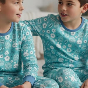May include: Two matching teal pajamas sets with an Easter-themed pattern of eggs, bunnies, and flowers. The long-sleeved tops and pants have blue trim. The pajamas are worn by two people.