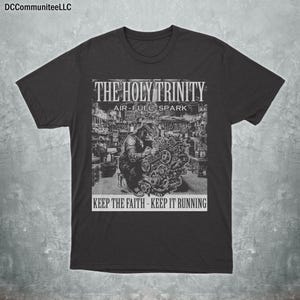 May include: Charcoal grey t-shirt with a black and white graphic. The graphic displays the text "THE HOLY TRINITY AIR-FUEL-SPARK" above an image of a person working on an engine. The text "KEEP THE FAITH - KEEP IT RUNNING" is below the image.