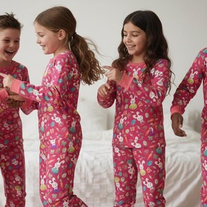 May include: Five children wearing matching pink pyjamas with an Easter-themed print of bunnies, carrots, and eggs. The pyjamas have long sleeves and trousers. The children are smiling and interacting.