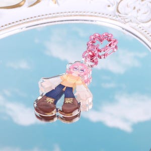 May include: A clear acrylic keychain featuring a cartoon character with pink hair, wearing glasses, a yellow shirt, blue jeans, and brown shoes. The keychain has a pink metal chain.