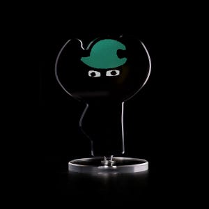 以下が含まれることがあります： 緑色の帽子をかぶった白い目のキャラクターの黒と緑の置物。置物は透明なプラスチック製の台座の上に立っています。