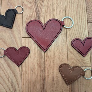 May include: Five leather heart-shaped keyrings with silver key rings. The keyrings are in different colours: black, red, brown, and two shades of burgundy.