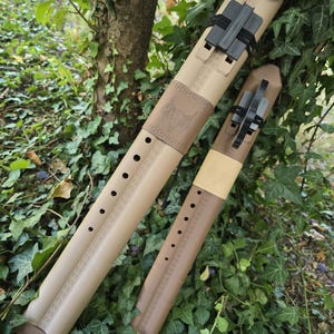 May include: Two tan and brown flute-like instruments with multiple finger holes and black clips. The larger instrument features an embossed bull head design. Both items have a cylindrical shape and are constructed from a smooth, solid material.