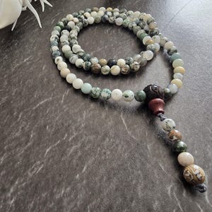 May include: A multi-coloured beaded necklace featuring white, green, and brown beads. The necklace has a decorative pendant with a carved design and a tassel of beads. The beads are round and appear to be made of stone.