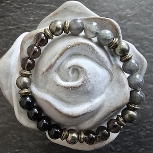 Peut inclure: Un bracelet de perles composé de perles gris foncé, noires et irisées. Il comprend des perles de quartz fumé, de labradorite et de pyrite, séparées par des entretoises couleur or antique. Le bracelet est présenté sur une sculpture en forme de rose blanche.