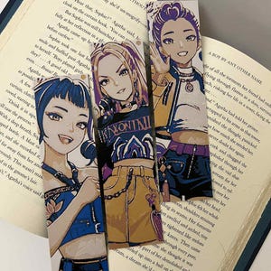 May include: Three anime-style bookmarks featuring illustrations of young women in stylish outfits. The bookmarks are set against the backdrop of an open book, with text visible on the pages. The characters have unique hairstyles and expressions.
