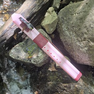 May include: A pink flute case with a dark red leather accent and a silver clasp. The case has a series of small, evenly spaced red dots. The flute case is resting on rocks near a stream.