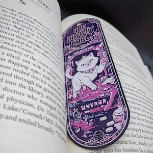 Puede incluir: Un marcapáginas rectangular con esquinas redondeadas que muestra un gato blanco con corona y collar. La ilustración en tonos morados y rosas incluye el texto "GOD DAMMIT CARL!" en la parte superior, un icono de gato en la parte inferior y varios objetos pequeños sobre un suelo de baldosas.