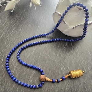May include: A beaded necklace featuring small, round, deep blue lapis lazuli stones. The necklace has a decorative gold-colored pendant and a wooden bead. The necklace is displayed on a dark gray surface.