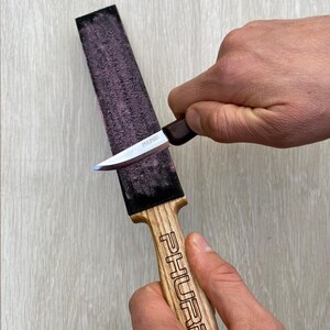 May include: A knife being sharpened on a whetstone. The whetstone is rectangular with a dark purple surface. The knife has a silver blade and a brown handle. The handle of the whetstone is wood-colored with the word "PHURE" engraved on it.