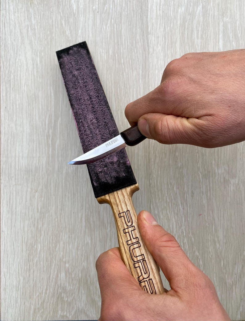May include: A knife being sharpened on a whetstone. The whetstone is rectangular with a dark purple surface. The knife has a silver blade and a brown handle. The handle of the whetstone is wood-colored with the word "PHURE" engraved on it.